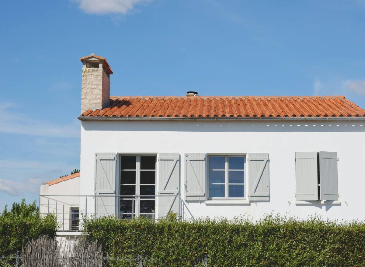 Maison individuelle blanche aux volets gris et au toit de tuiles, derrière une haie